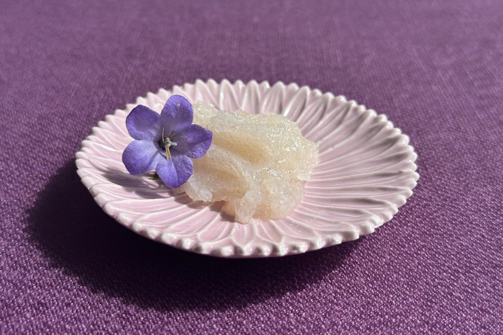 Kristallines Ziaja Cupuaçu Zuckerpeeling auf einem rosa Porzellanteller mit Glockenblumenblüte – sanftes Peeling mit Sheabutter und Macadamia-Öl