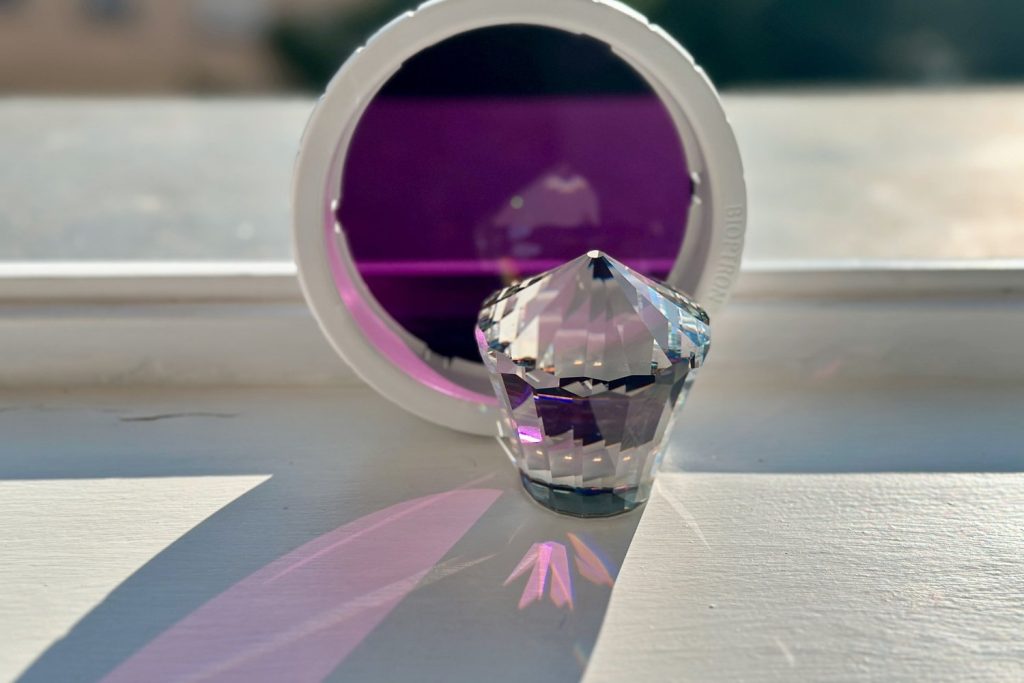 Bioptron-Violett-Aufsatz mit Bleikristall vor dem Fenster – warme Lichtbrechung in Rosa- und Violetttönen