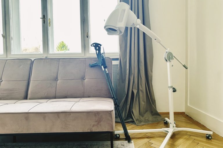 Eine Krücke lehnt am rosa Velvet-Sofa, daneben steht die Bioptron PRO 1 auf dem Standstativ — typische Anwendungssituation bei Sportverletzungen im Wohnzimmer