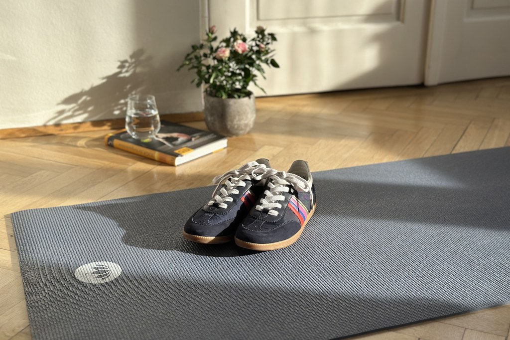 Dunkelblaue Sneakers auf einer grauen Yogamatte, daneben ein Glas Wasser, ein Yoga-Buch und ein Rosenstrauß im Sonnenlicht — bereit für die Rückkehr in die Bewegung.