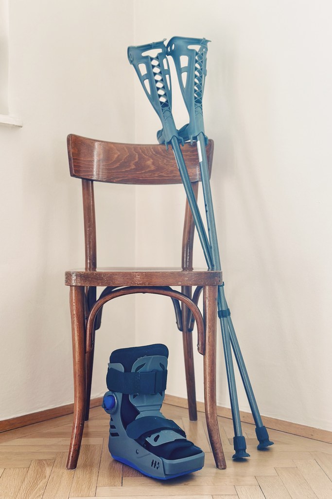 Ein Wiener Bugholzstuhl, auf dessen Lehne zwei blaugraue Krücken lehnen, daneben am Boden ein orthopädischer Stiefel — Hilfsmittel nach einer Knöchel-Operation.