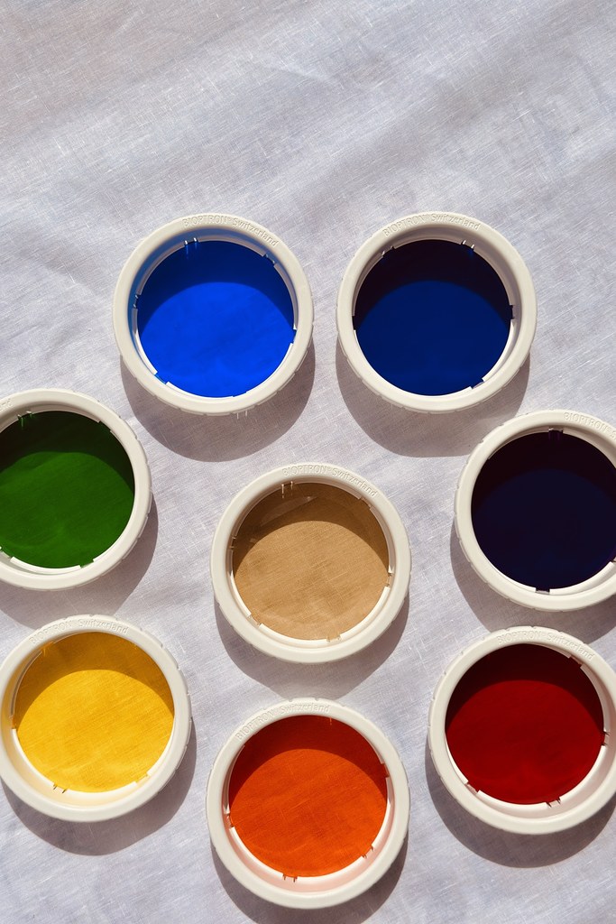 Acht Bioptron-Farbaufsätze in den Farben des Lichtspektrums — Rot, Orange, Gelb, Grün, Blau, Indigo, Violett und das weiße Hyperlicht in der Mitte, auf hellem Leinen arrangiert
