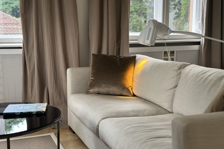 Bioptron-Lampe auf einem hellen Sofa mit samtigem Kissen — der warme Lichtkegel der Lampe fällt auf das Kissen und schafft eine ruhige Atmosphäre für die Lichttherapie zu Hause.