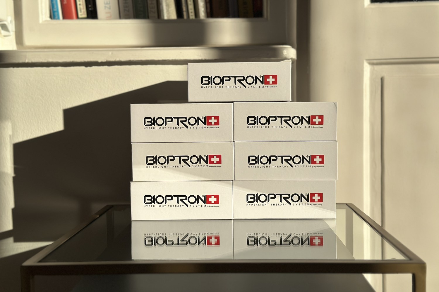 Acht weiße Bioptron-Verpackungen, sauber zu einer Pyramide gestapelt auf einem Glastisch, im Hintergrund eine Bücherwand im warmen Sonnenlicht — Beweis langjähriger eigener Erfahrung mit der Bioptron-Lichttherapie.