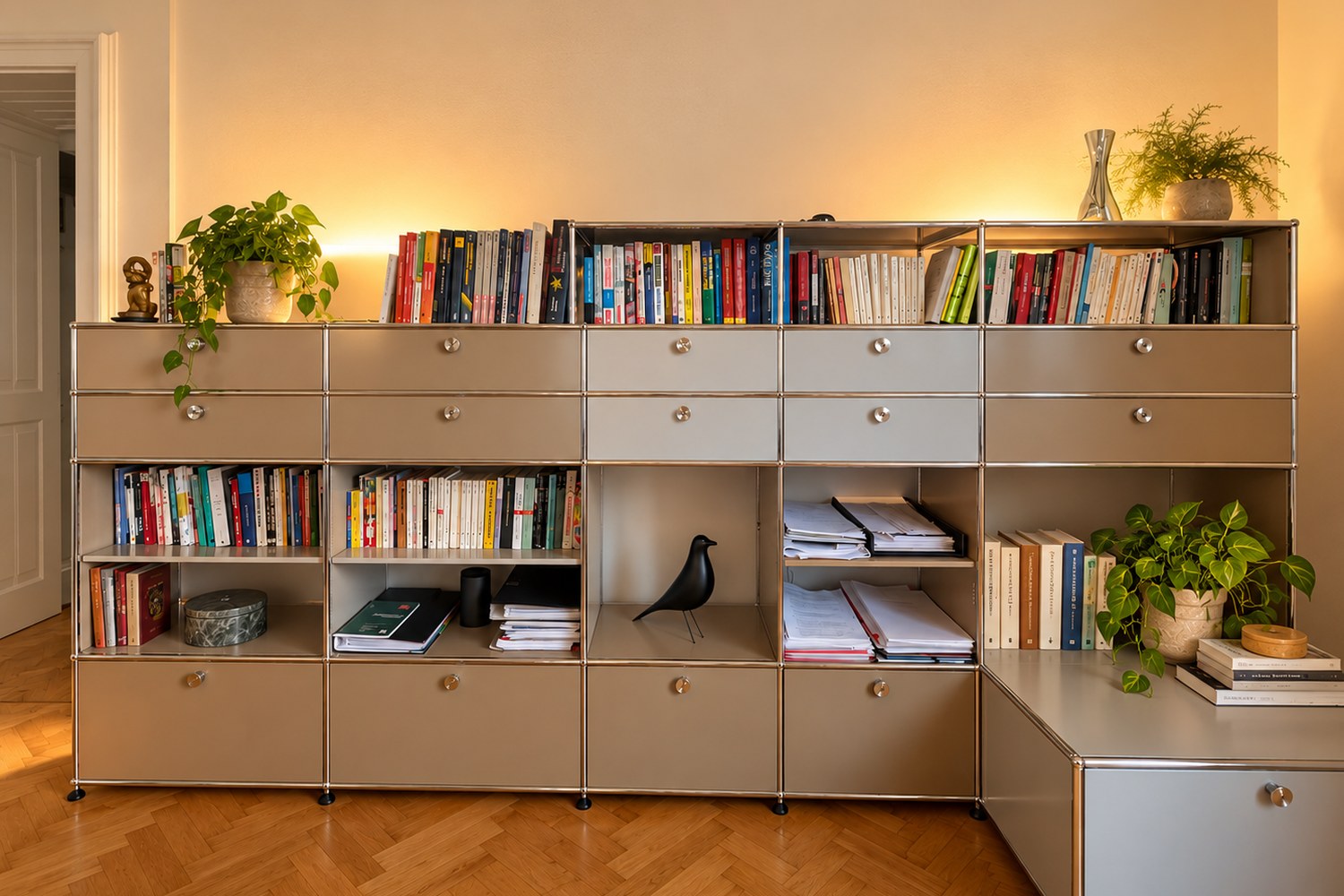 Ein zweifarbiges Metallregal in Grau und Braun mit Schubladen und offenen Fächern voller Bücher, dekoriert mit Grünpflanzen und einem schwarzen Holzvogel, in warmes Abendlicht getaucht — mein persönliches Bücherregal mit Lektüre rund um Bewusstsein und innere Arbeit.