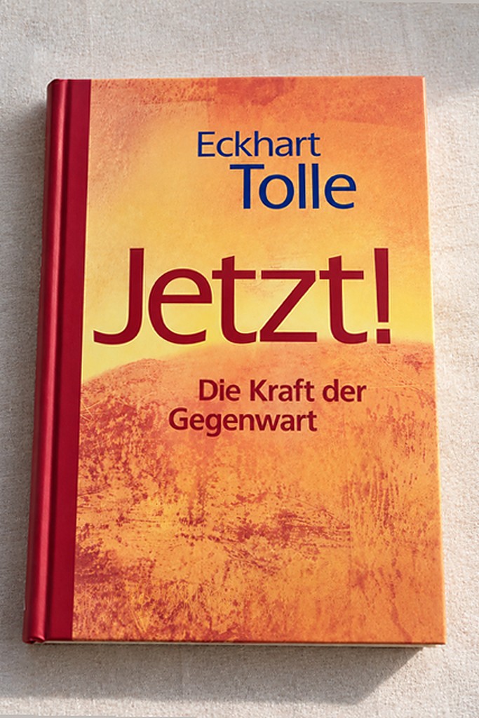 Das Buch „Jetzt! Die Kraft der Gegenwart" von Eckhart Tolle mit orange-gelbem Cover und rotem Buchrücken, liegend auf hellem Leinen fotografiert.