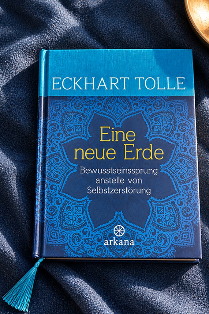 Das Buch „Eine neue Erde" von Eckhart Tolle mit blauem Mandala-Cover und türkiser Lesezeichen-Quaste, liegend auf dunklem Stoff neben einer Klangschale.