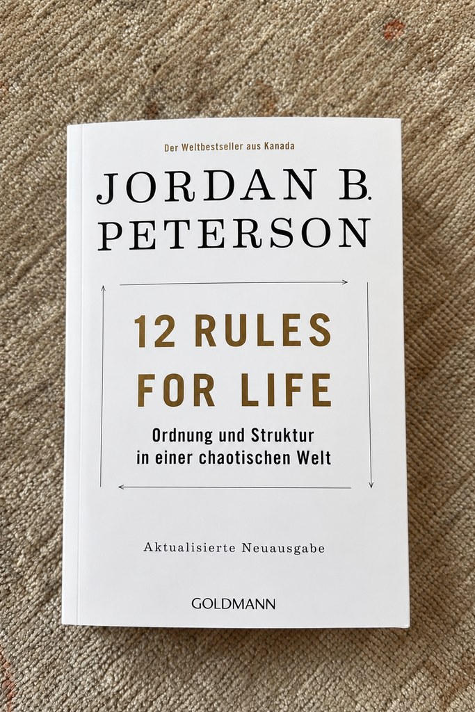 Das Taschenbuch „12 Rules for Life — Ordnung und Struktur in einer chaotischen Welt" von Jordan B. Peterson mit weißem Cover, liegend auf einem beigen Teppich.