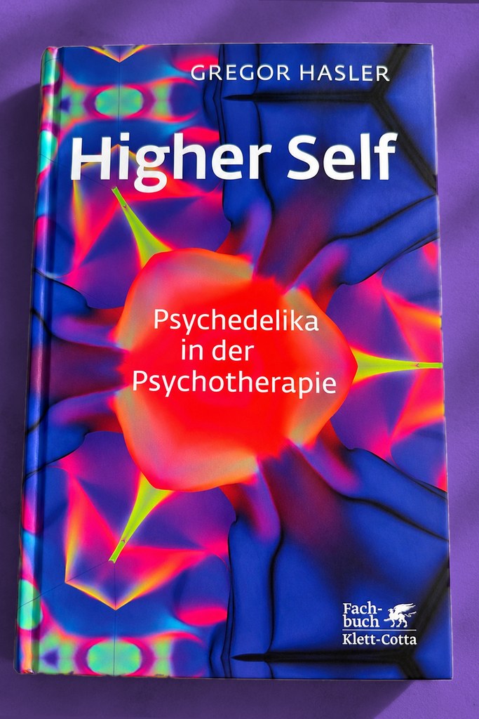 Das Buch „Higher Self — Psychedelika in der Psychotherapie" von Gregor Hasler mit leuchtend blau-rotem, kaleidoskopartigem Cover, liegend auf violettem Untergrund im Sonnenlicht.