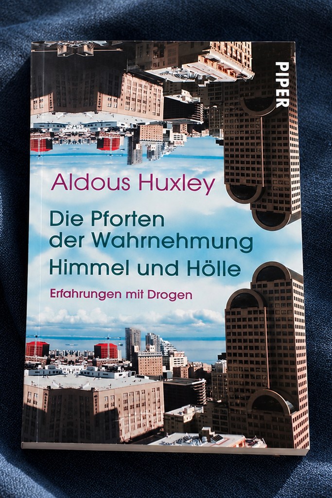 Das Taschenbuch „Die Pforten der Wahrnehmung / Himmel und Hölle" von Aldous Huxley mit einem Cover aus gespiegelten Hochhäusern und Himmel, liegend auf dunkelblauem Stoff.